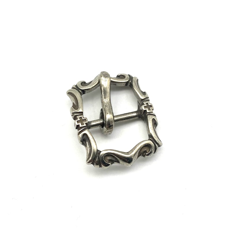 May include: Silver-toned metal belt buckle with an ornate design. The buckle has a square shape with rounded corners and intricate details.