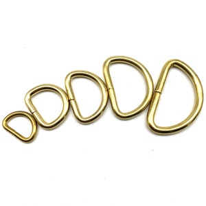 May include: Five gold-colored metal D-rings of varying sizes. The rings are arranged in a semi-circle, with the smallest ring at the bottom and the largest ring at the top.
