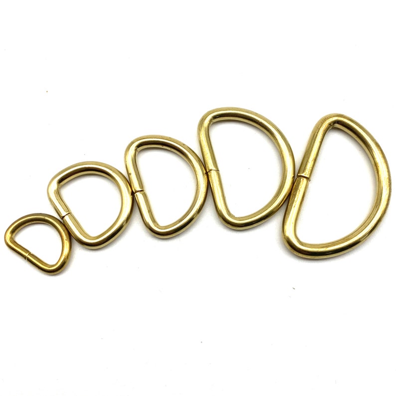 May include: Five gold-colored metal D-rings of varying sizes. The rings are arranged in a semi-circle, with the smallest ring at the bottom and the largest ring at the top.