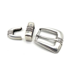 May include: A silver belt buckle set with a decorative design. The set includes a buckle, a keeper, and a prong. The buckle is a traditional style with a smooth, rounded edge. The keeper and prong are both simple and functional.