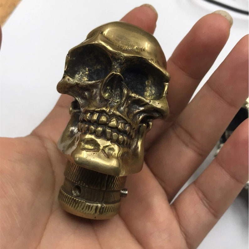 May include: A gold-colored skull-shaped knob with a detailed design. The knob has a threaded base for attaching to a surface.