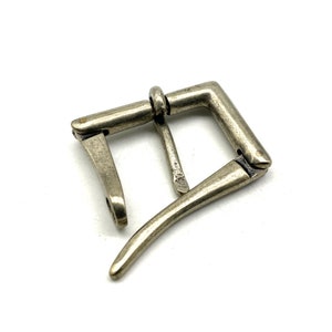 Könnte beinhalten: Silberfarbener Metallschnalle mit einem D-Ring und einem Federschnappverschluss. Die Schnalle ist für die Verwendung mit einem Riemen oder Gürtel konzipiert.