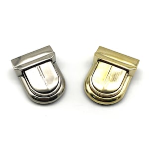Pode incluir: Dois fechos metálicos, um prateado e outro dourado, com uma parte superior arredondada e arqueada e uma ranhura decorativa.