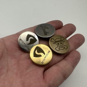 Könnte beinhalten: Eine Sammlung runder Metallmarken in verschiedenen Farben, darunter Silber, Gold und Bronze. Einige Marken zeigen den Text "Linda", ein Storchenlogo und ein Totenkopfdesign. Die Marken werden in einer Hand gehalten.