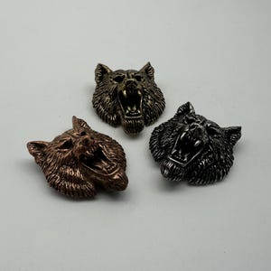 Può includere: Tre sculture di teste di lupo in metallo nei toni bronzo, argento e oro. Ogni testa di lupo è scolpita con la bocca aperta, mostrando i denti. Le sculture sono dettagliate con pelliccia strutturata e su sfondo bianco.