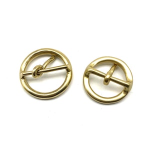 May include: Two gold-toned metal belt buckles. One buckle has a knotted bar, the other has a straight bar.