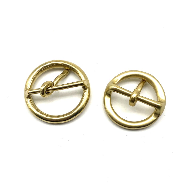 May include: Two gold-toned metal belt buckles. One buckle has a knotted bar, the other has a straight bar.