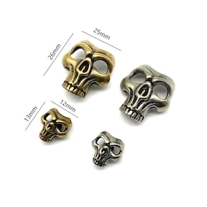 Könnte beinhalten: Vier Totenkopf-Anhänger aus Metall in verschiedenen Größen. Der größte Anhänger ist 25 mm breit und 26 mm hoch, der zweitgrößte ist 13 mm breit und 12 mm hoch, und die beiden kleinsten sind 13 mm breit und 12 mm hoch. Die Anhänger sind silber und goldfarben.