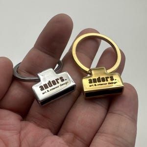 Può includere: Sono mostrati due portachiavi, uno argentato e uno dorato. Ogni portachiavi presenta un'etichetta rettangolare con la scritta "anders. art & interior design" in un carattere marrone scuro. Gli anelli portachiavi sono circolari. Il portachiavi argentato ha un anello argentato e il portachiavi dorato ha un anello dorato.