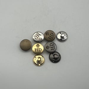 Pode incluir: Uma coleção de botões de pressão de metal em vários acabamentos, incluindo prata, ouro e bronze. Cada botão apresenta as letras "HD" e a palavra "Handmade". Estes botões são ideais para adicionar um toque personalizado a roupas e acessórios.