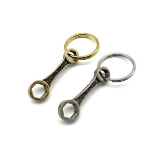 Puede incluir: Dos llaveros con forma de llave inglesa, uno dorado y otro plateado, con el texto "POTONG METAL" grabado en cada uno.