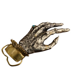 May include: A silver hand with a turquoise stone ring, the hand is holding a gold belt buckle with a decorative design.