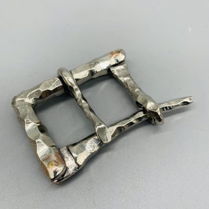 Puede incluir: Una hebilla de metal plateado con textura martillada. La hebilla tiene una forma rectangular con una abertura redondeada para un cinturón.