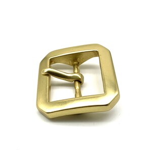 May include: A gold-toned metal buckle with a rectangular shape and a smooth, polished finish. The buckle has a single prong and a curved tongue.
