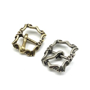 May include: Two ornate metal belt buckles, one in gold and one in silver, with a decorative design and a cross pattern.