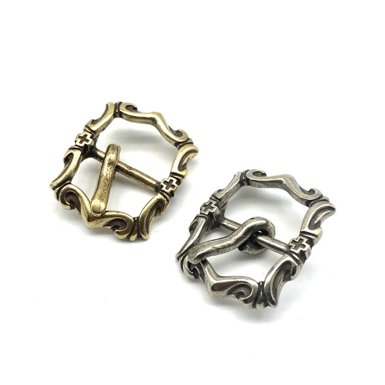 May include: Two ornate metal belt buckles, one in gold and one in silver, with a decorative design and a cross pattern.