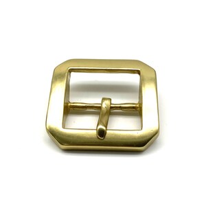 May include: A gold-toned metal buckle with a rectangular shape and a smooth finish. The buckle has a single prong for attaching to a strap.