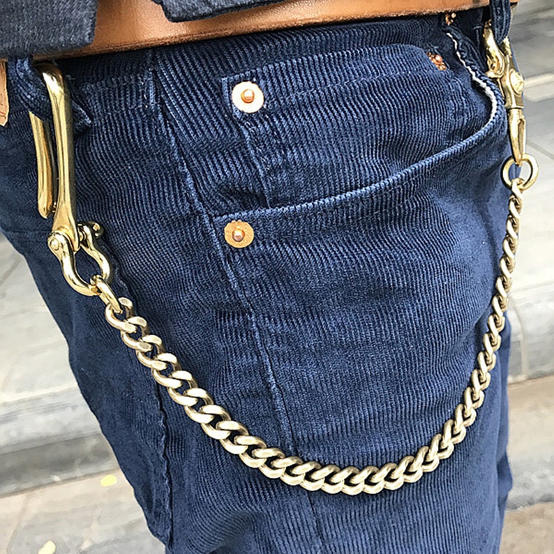 May include: A gold-colored chain wallet accessory attached to a pair of navy blue corduroy pants. The chain is thick and has a hook on each end. The pants have a brown leather belt and gold-colored rivets.