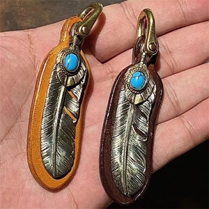 Peut inclure: Deux porte-clés en cuir, un beige et un marron, chacun avec un motif de plume argentée. Une pierre turquoise est sertie dans un métal couleur laiton en haut, avec un fermoir en laiton. Les porte-clés sont conçus pour être attachés à des clés ou des sacs.