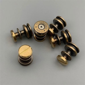 Puede incluir: Un conjunto de siete cierres metálicos de color latón. Cada cierre tiene una cabeza redonda con un borde elevado y un poste roscado. Uno de los cierres tiene una cabeza redonda con las palabras "PT. HANDMADE 120G" grabadas en él.