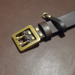 May include: A brown leather belt with a brass buckle and a loop on the end. The buckle has a double-prong design.