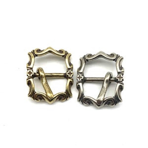 May include: Two ornate metal belt buckles, one gold and one silver, with a decorative design. Both buckles have a square shape with a curved top and a cross design on each side.