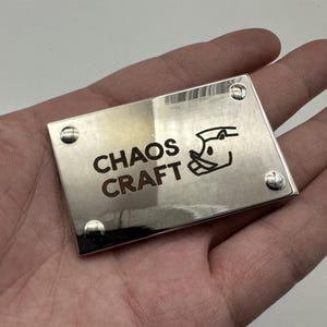 Pode incluir: Placa metálica retangular prateada com cantos arredondados e quatro rebites. As palavras "CHAOS CRAFT" estão gravadas em cor escura, juntamente com um gráfico estilizado. A placa é segurada em uma mão.