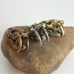 May include: Three metal animal skull rings with detailed carvings of teeth and fangs. The rings are in gold, silver, and bronze finishes.
