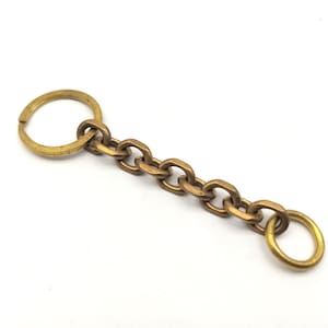 Puede incluir: Un llavero dorado con un anillo redondo en un extremo y un anillo redondo más pequeño en el otro extremo. La cadena está hecha de eslabones ovalados.