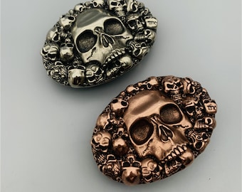 May include: Two silver and copper belt buckles with a skull design. The buckles are surrounded by smaller skulls.