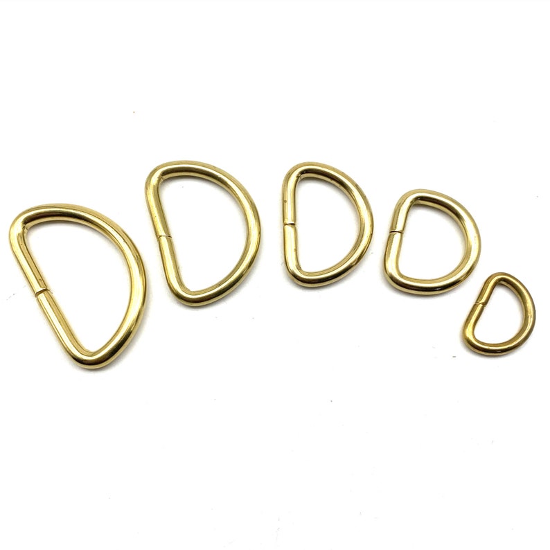 May include: Five gold-colored metal D-rings of varying sizes. The rings are arranged in a semi-circle on a white background.