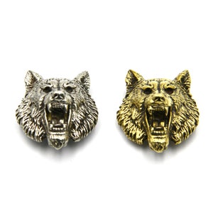 Könnte beinhalten: Zwei Metallknöpfe in Form eines Wolfskopfes, einer silberfarben und einer goldfarben, mit einem knurrenden Ausdruck.