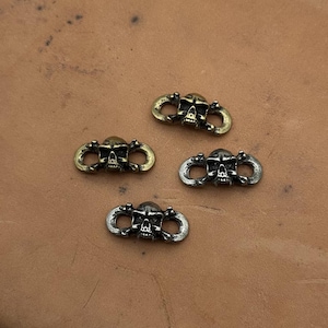 Puede incluir: Cuatro cierres metálicos con forma de calavera en tonos dorados y plateados. Cada cierre presenta un diseño detallado de calavera con un lazo a cada lado. Los cierres están dispuestos sobre una superficie marrón texturizada.