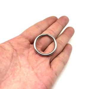 Puede incluir: Un anillo metálico plateado y brillante de forma circular. El anillo se sostiene en la palma de una mano, y el fondo es blanco.