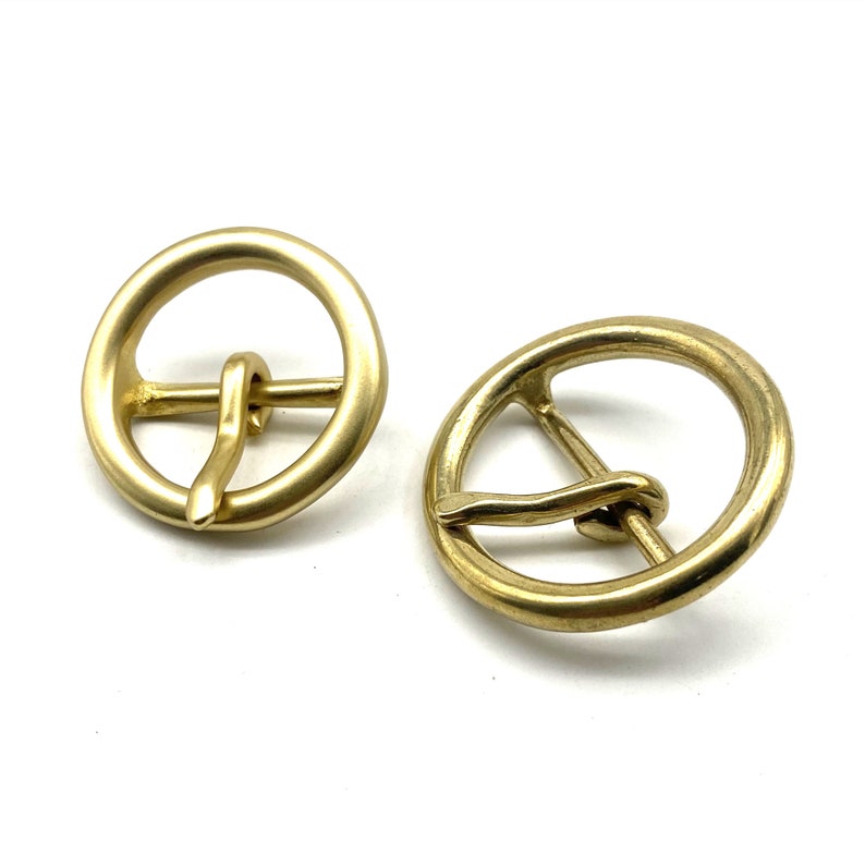 May include: Two gold-toned metal belt buckles with a round shape and a simple design. The buckles have a smooth, polished finish.