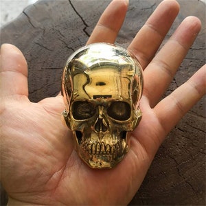 Puede incluir: Una escultura de calavera de metal de color dorado. La calavera está detallada con un acabado liso y brillante y presenta rasgos faciales realistas.