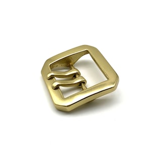 May include: A gold-toned metal buckle with a rectangular shape and three prongs. The buckle is designed for a belt or strap.