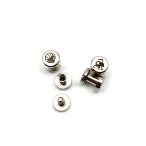 Op de afbeelding: Een verzameling zilverkleurige metalen klinknagels met gegraveerde ontwerpen. Sommige klinknagels hebben een schelpmotief, andere een gestileerd dier. De klinknagels zijn verspreid over een witte ondergrond.