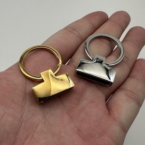 Puede incluir: Dos piezas de herrajes para llaveros metálicos. Una es dorada, la otra plateada. Cada una tiene una base rectangular con un lazo para sujetar las llaves. Los herrajes se muestran sobre un fondo neutro.