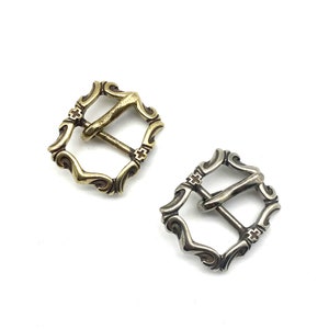 May include: Two ornate metal belt buckles, one gold and one silver, with a cross design. The buckles are square shaped with intricate scrollwork.