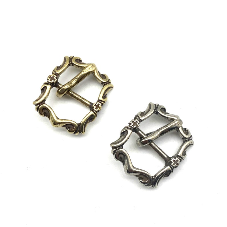 May include: Two ornate metal belt buckles, one gold and one silver, with a cross design. The buckles are square shaped with intricate scrollwork.