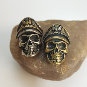 Puede incluir: Dos anillos de calavera de plata con detalles dorados. Cada calavera lleva un sombrero de estilo militar con un emblema de águila. Los anillos están sobre una roca marrón.