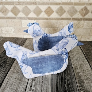 May include: Two fabric chicken-shaped kitchen accessories featuring a white base with blue illustrations of roosters, hens, and baskets of eggs. Each chicken has a solid blue denim-like wing patch and a small blue and white checkered comb.