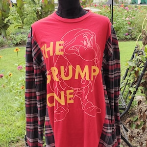 May include: A long-sleeve shirt with a red front panel featuring a yellow graphic of the character Grumpy and the text THE GRUMPY ONE. The sleeves and back are made of a black, red, and grey plaid fabric with a crew neckline and buttoned cuffs.