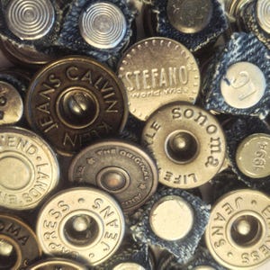 May include: A collection of assorted metal jean buttons, some with denim backing. The buttons feature brand names like "Calvin Klein Jeans", "Stefano", and "Sonoma Jeans". The buttons are in various shades of silver and bronze.