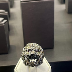 Può includere: Un anello dettagliato con un design a testa di leone. L'anello ha una base in metallo scuro con una criniera strutturata e piccole pietre scintillanti. Il leone ha occhi rossi e la bocca aperta, mostrando i suoi denti.