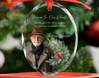 Op de afbeelding: Ovale glazen kerstversiering met een rood lint. De versiering toont een foto van een man in een bruine jas en hoed, met de tekst "Forever In Our Hearts" en "Thomas Grace 1968-2025".