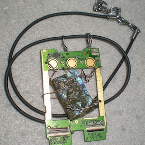 May include: A necklace featuring a rectangular green circuit board pendant with three gold circular contact points. A rectangular, iridescent, dark blue and green stone is secured to the center of the board with thin, bent copper-colored wire.