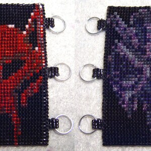 以下が含まれることがあります： 黒、赤、紫のビーズでできたキーホルダーが2つ。左側のキーホルダーは赤い悪魔のデザイン。右側のキーホルダーは紫の花のデザインです。