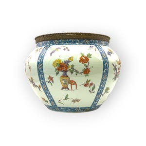 May include: A white ceramic planter with a bronze rim and blue floral accents. The planter features a detailed design of flowers, birds, and a vase with a small table. The planter is a decorative piece.